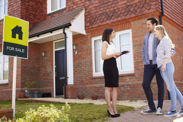 Couple standing in front of a house with a "For Sale" sign.