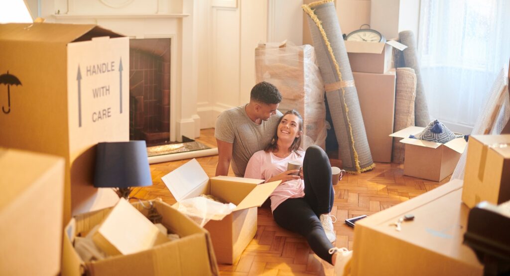 A young couple sitting on the floor surrounded by moving boxes, relaxing and unpacking in their new home — ideal for illustrating a move to Tonbridge, Kent.