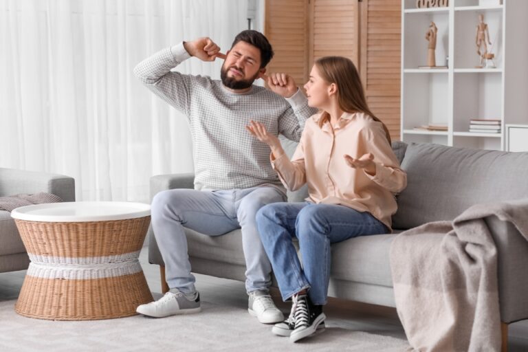 A man and woman sit on a couch, frustrated by noisy neighbours in their living room.