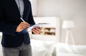 A man in a suit stands by a couch, holding a clipboard and looking thoughtfully at the space around him.