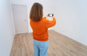 A person in an orange sweater and jeans takes a photo with a smartphone in an empty room with wooden floors and white walls.