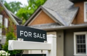 A for sale sign stands in front of a house, indicating the property is available for purchase.