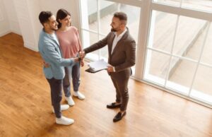 A couple stands in an empty room while shaking hands with a man in a suit holding a clipboard, likely concluding a real estate transaction.