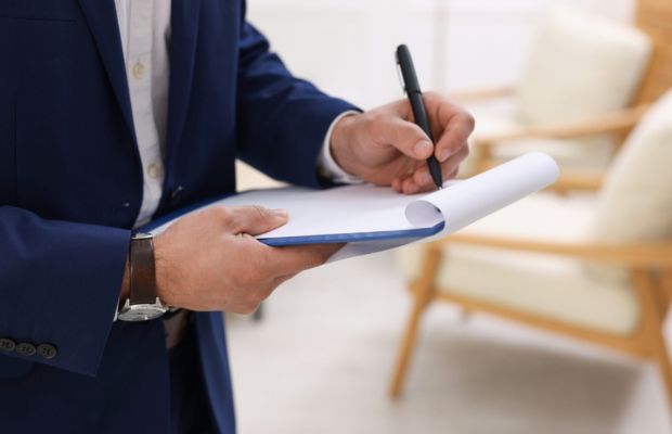 A person in a blue suit is writing on a clipboard with a pen, standing in a room with chairs in the background.