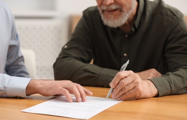 Two people sit at a table; one points to a document while the other holds a pen, appearing to review or sign the paperwork.