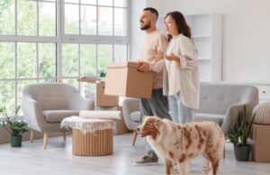 A couple carries boxes into their new home while their dog excitedly explores the front yard.
