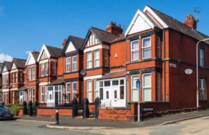 A row of red-brick Victorian terraced houses with white trim and steep gables lines a sunny street, creating a bright, welcoming neighborhood scene.
