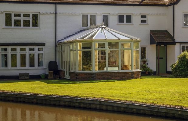 A white house with a glass conservatory extension overlooking a grass lawn and a canal.